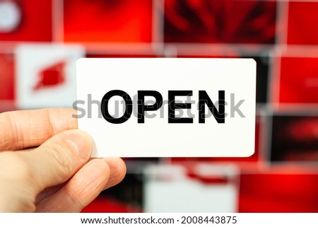 A man on a red background holds a business card with the word - Open. Say "Welcome". We are open.