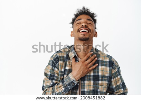 Close up portrait of happy, relieved and carefree african american guy 20s years, laughing out loud, raise head up with closed eyes and big cheerful smile, holding hand on chest, white background Royalty-Free Stock Photo #2008451882