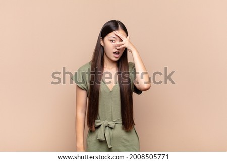 asian young woman looking shocked, scared or terrified, covering face with hand and peeking between fingers
