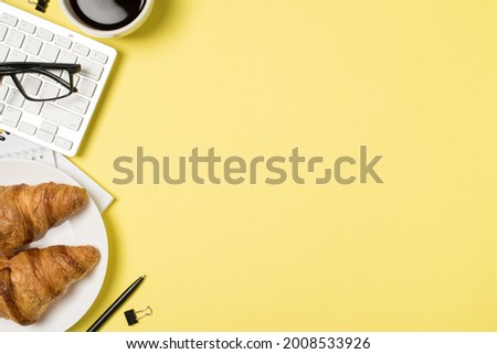 Top view photo of workplace glasses on keyboard cup of coffee notebooks binder clip pen and plate with croissants on isolated pastel yellow background with copyspace