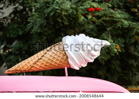 A large figure is a sign on the van.White vanilla ice cream in a waffle cone.Food court with fast food on the street against the background of green tree branches in the park.Cold sweet dessert.