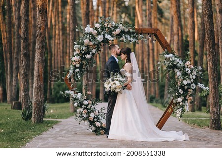 Stylish groom in a suit and a cute brunette bride in a white dress in the forest near a wedding wooden arch decorated with flowers. Wedding portrait of the newlyweds. Royalty-Free Stock Photo #2008551338