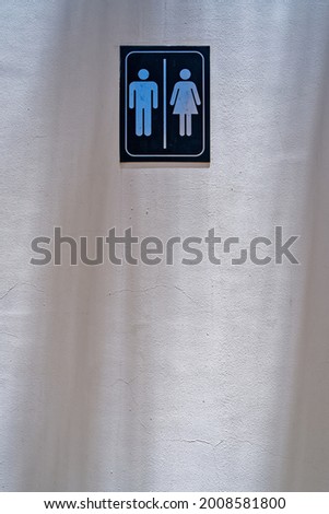A sign with the designation of a public men's and women's toilet is located on a textured wall with shadows from palm trees near the beach or pool. Human silhouettes are printed on a black plate.