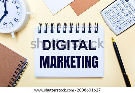 On a light background, a white alarm clock, a calculator, a pen and a notebook with the text DIGITAL MARKETING. Flat lay