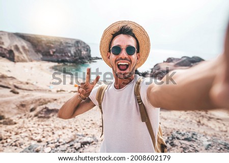 Young man with backpack taking selfie portrait on a mountain - Smiling happy guy enjoying summer holidays at the beach - Millennial showing victory hands symbol to the camera - Youth and journey  Royalty-Free Stock Photo #2008602179