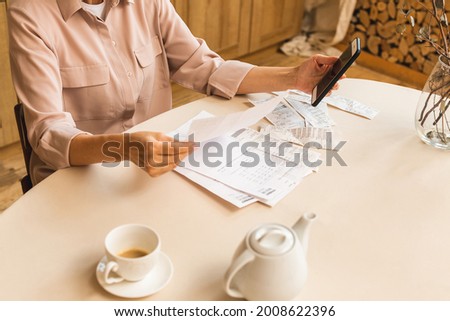 Middle aged mature senior woman holding paper bill or letter at home for making online payments on website on mobile phone, calculating financial taxes fee cost, reviewing bank account. Royalty-Free Stock Photo #2008622396