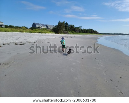 A young boy walking along water at a Nova Scotian beach. She's walking her full breed, black and tan Dachshund on a leash while holding her shoes in her other hand. The waves are rolling in.