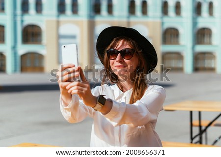 Caucasian girl in a hat and sunglasses smiling while sitting in a cafe in the summer and takes pictures on a smartphone.