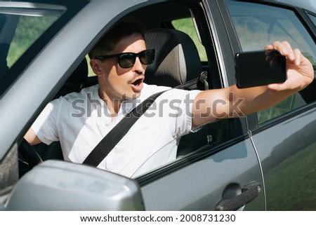 Emotional portrait of man driver with glasses and with open mouth taking pictures on mobile phone in an open window while driving a car. Surprise concept on the road, incredible incident, wow effect