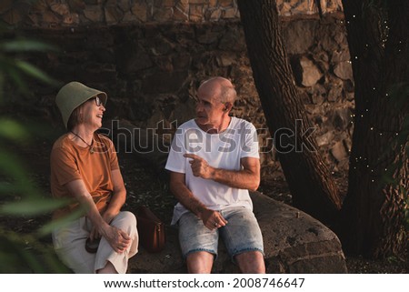 Happy senior couple (Asian woman and Caucasian man) sitting in tree shadow in city park and having a talk and laughing in Summer day