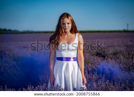 young beautiful attractive girl in white dress is looking in camera in lavender field with smoke on background during sunrise