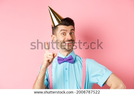 Holidays and celebration concept. Image of happy man in birthday party hat pitching an idea, raising finger and smiling, have plan or solution, standing over pink background