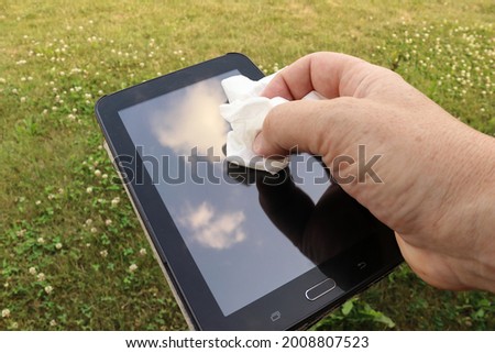 Cleaning the screen of an old tablet. A blue sky with white clouds can be seen in the reflection of the display. Green grass in the background.
