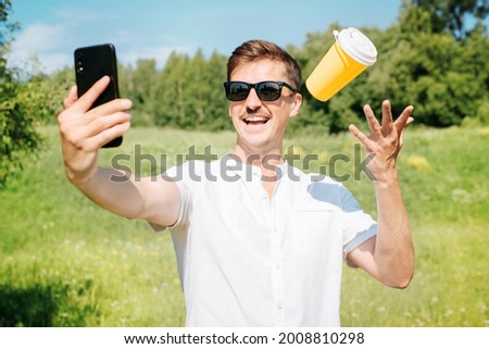 Happy man wearing glasses tossing disposable cup of coffee and taking selfie on mobile phone, outdoors summer day. Portrait of smiling mustachioed young man rejoicing at phone camera outside in park