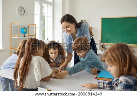 Happy school teacher and students having interesting class in modern classroom. Little children reading book, looking at pictures, learning, discussing new things, working on creative project together