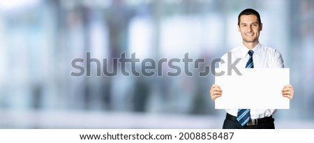 Happy smiling businessman in white shirt and tie, showing blank signboard with copy space area for text or sign, blurred modern office background. Confident business man holding paper board. Mock up