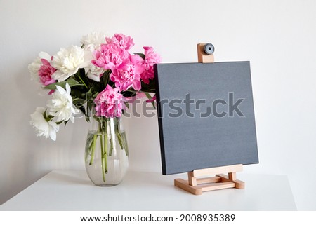 Black canvas mockup on wooden easel and vase with pink flowers on table on white wall background. Blank artistic canvas