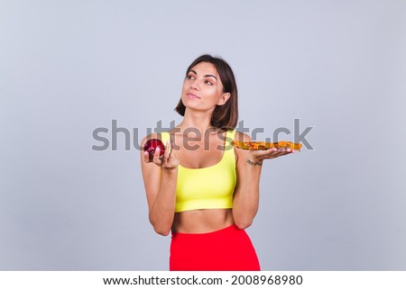 Sports woman stands on gray background, satisfied with the results of fitness training and diet, holds apple and pizza in hands thoughtful