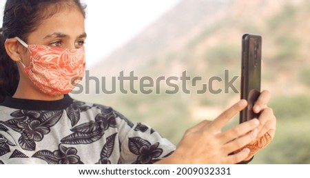 A shallow focus of an Indian girl wearing a face mask and taking a picture on her smartphone
