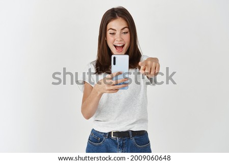 Excited young woman taking picture on mobile phone, pointing at camera and smiling, recording video on smartphone, white background