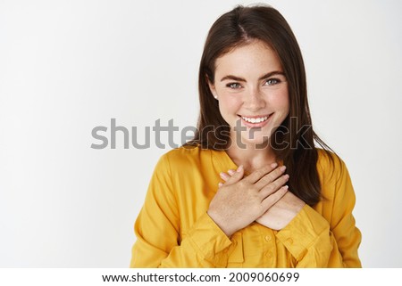 Close-up of pretty female thanking you, holding hands on heart and smiling delighted, standing over white background