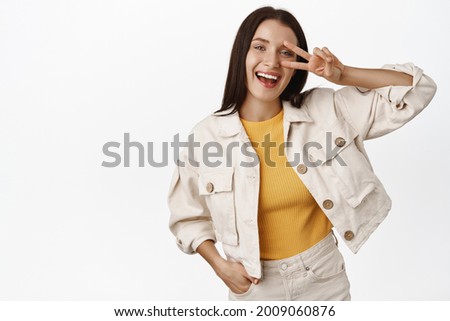 Positive adult woman laughing, smiling happy and showing peace v-sign gesture near eye, stay optimistic, standing against white background
