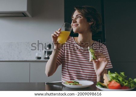 Young housewife woman 20s in casual clothes striped t-shirt eat vegeterian breakfast lunch drinking orange juice eat sandwich cooking food in light kitchen at home alone Healthy diet lifestyle concept Royalty-Free Stock Photo #2009098616