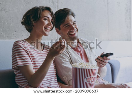 Side view young cheerful couple two friend woman man 20s wearing casual clothes sit on sofa hold takeaway bucket eat popcron watch movie rest indoors at home flat together. People lifestyle concept. Royalty-Free Stock Photo #2009098637