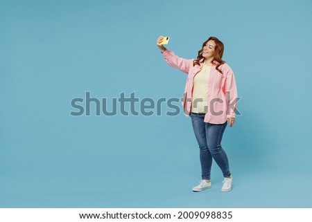 Full length young redhead chubby overweight woman 30s with curly hair wear pink shirt casual clothes doing selfie shot on mobile phone post photo on social network isolated on pastel blue background