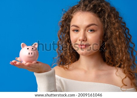 Curly-haired young woman holding piggy bank against blue background