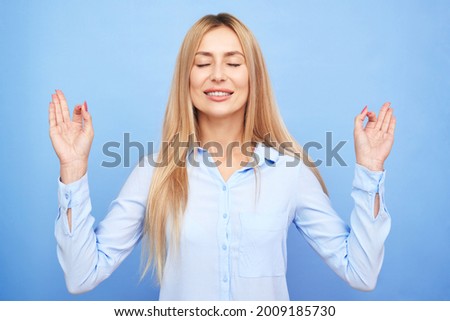 Businesswoman in a blue shirt keeps eyes closed holding fingers in mudra gesture, beautiful manager girl keeps calm relaxes isolated on blue background, anti-stress therapy meditation