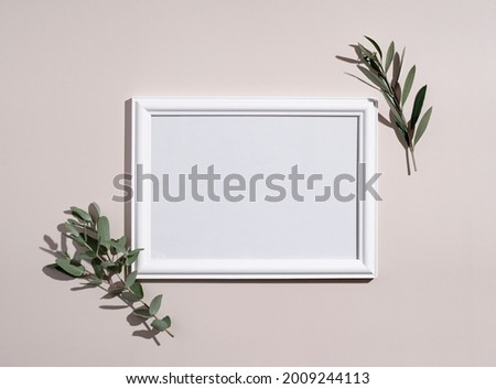Picture frame mockup with white wooden frame. Eucalyptus leaves on beige background. Natural light and shadow overlay. Flat lay, top view.