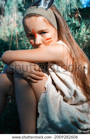 Indian girl with red makeup on her face posing in green grass. Portrait of little cute girl with long hair in style of the American Indians