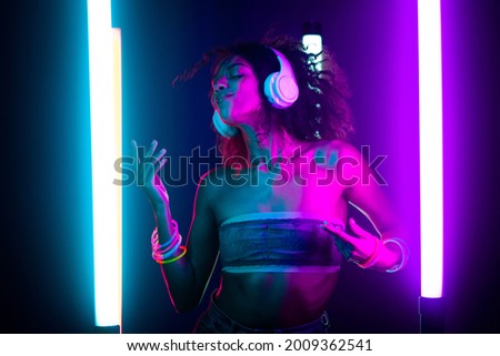 Portrait of party girl dancing to music with headphones on glowing multi-colored lamps in studio. Disco woman with curly hairstyle, modern outfit. Amazing style. Royalty-Free Stock Photo #2009362541