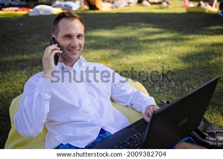Handsome Man Sitting On The Grass In The City With A Laptop And Talking On Phone