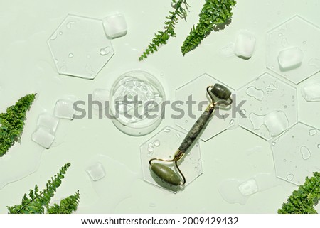 Self made moisturizer and green jade face roller with ice cubes. Exotic fern leaves on mint green background. Minimal flat lay, top view. Facial massage concept, handmade cosmetics.