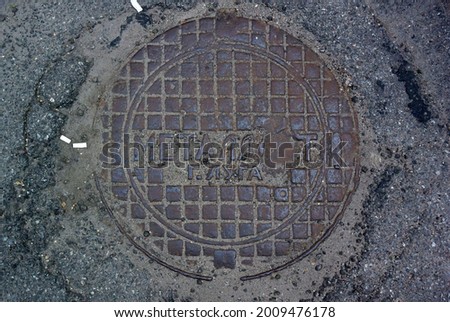 Photo of a background with a metal round sewer hatch pattern of squares and a Russian lettering. Translated from Russian: Metalist, city of Luga.
