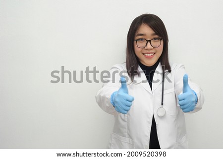 Asian female young doctor showing thumb up hand sign with glasses wearing white coat with stethoscope in white clean background.