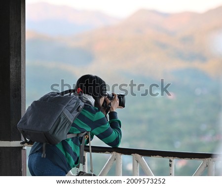 An Asian female tourist wearing a green striped long-sleeve shirt with a blue backpack is photographing the mist-shrouded nature. concept of vacation travel