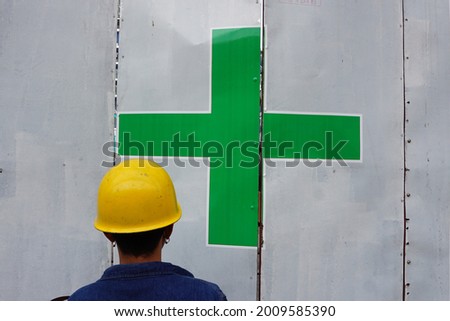 The back of a man wearing a yellow safety helmet, blue denim jacket and round metal earrings on both sides. He stood in front of a large white metal plate marked green cross. Work safety concept.