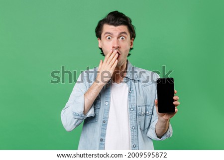 Shocked young man 20s in casual clothes white t-shirt denim shirt hold mobile phone with blank empty screen mock up copy space covering mouth with hand isolated on green background studio portrait