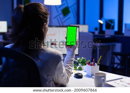 Freelancer looking at mobile phone with green screen display during online videocall meeting sitting at desk in business office. Businesswoman watching desktop monitor with green mockup, chroma key