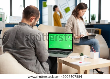 Over the sholder of manager sitting on couch holding laptop with green screen, mockup desktop while diverse team working on background. Multiethnic people planning project on chroma key display