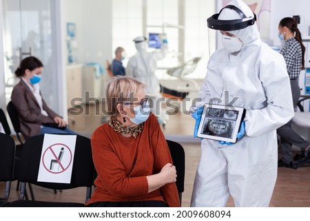 Dentist doctor with face shield pointing on tablet display explaining dental x-ray to senior patient during global pandemic. Nurse wearing protection suit, overall, mask and gloves, new normal.