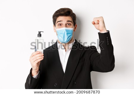 Concept of covid-19, business and social distancing. Close-up of happy man in trendy suit and medical mask, cheering and raising hand up, showing hand sanitizer, white background
