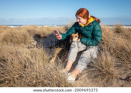 Young red haired girl taking a selfie of her and her pet dog, on a clear blue winter's day on sand dunes at a remote beach, near Gisborne, New Zealand