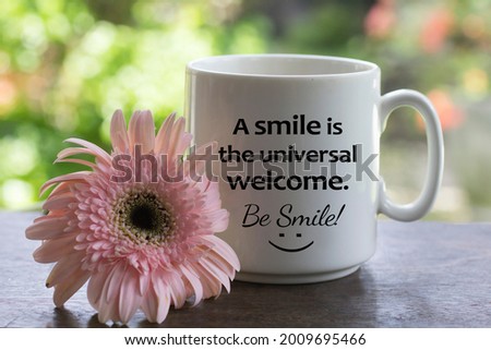 Positive message on a white coffee cup - A smile is the universal welcome. Be smile! with a happy smiling sign on it and soft pink daisy flower on the table. Kindness begin with a smile concept.