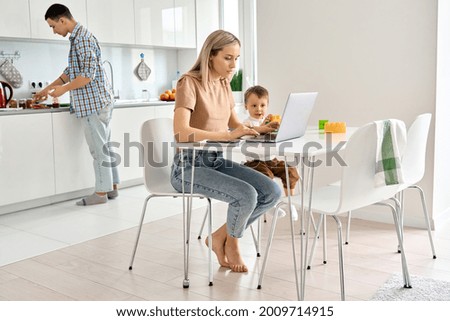 Busy young mother working from home office using laptop computer sitting at kitchen table with cute kid child son at home while dad cooking. Parents and child, distance virtual work in family life.