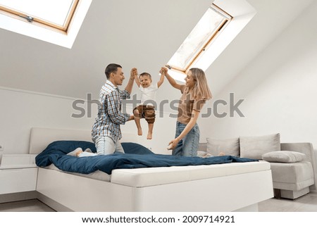 Happy young family couple playing with cute small toddler kid son in bedroom at home. Parents and child boy having fun feeling joy jumping on bed bonding spending weekend morning time together.