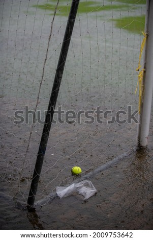 A flood softball pitch full of water with a green ball left nearby the soaking area on a raining field. Viewers waiting for a league match to begin while the field fulls with water without any player.